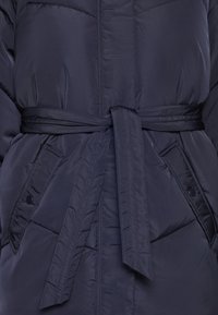 Námořnické prošívané sako s uvázaným páskem v pase, obsahující dvě přední kapsy a podšívku s prošíváním. Hladký povrch bez viditelného hardware.