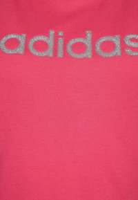 Camiseta de algodón rosa con el logo brillante en plata de "adidas" en el pecho. Textura suave, cuello redondo, mangas cortas.