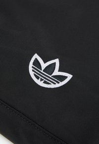 Černá látka s vyšitým černým logem Adidas trefoil obrysovaným bílou barvou poblíž šitého švu.