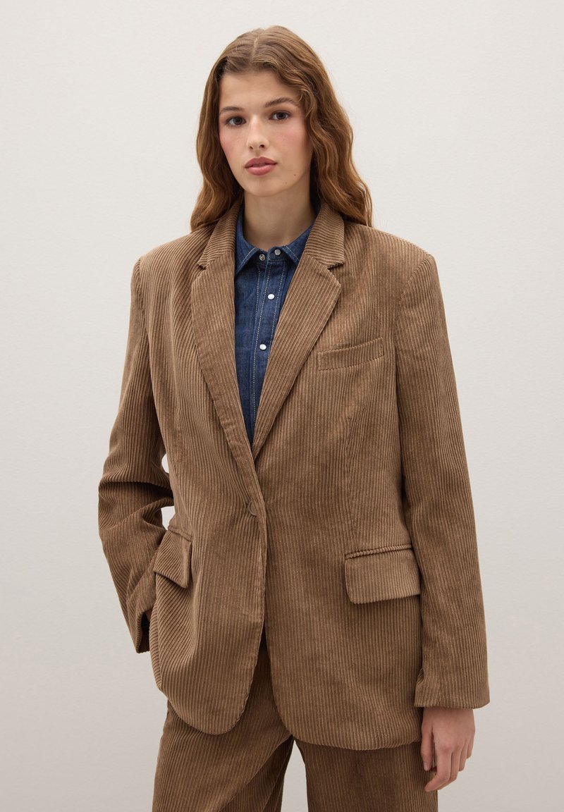 Bruine corduroy blazer met een notch lapel, twee voorzakken en verticale strepen, gelaagd over een blauw denim shirt met knopen.