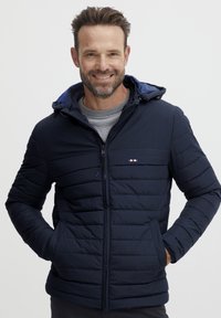 Jachetă puffer bleumarin cu glugă, având un design cu quilt orizontal și închidere cu fermoar. Două buzunare laterale și detalii accentuate în zona pieptului.
