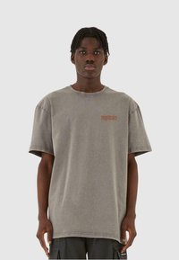 Grå oversized bomulls T-skjorte med korte ermer, med en subtil logo i rødbrun på venstre side og rund hals.