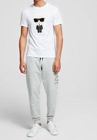 Camiseta de algodón blanca con un gráfico de personaje de dibujos animados en blanco y negro. Combinada con pantalones de chándal grises con impresión de logo y zapatillas blancas.