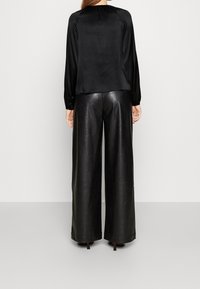 Donna con una blusa in satin nera a maniche lunghe e pantaloni in pelle neri a gamba larga e vita alta, in piedi contro uno sfondo grigio uniforme.