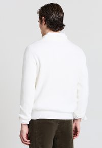 Maglione bianco a coste con colletto alto e maniche lunghe. Il tessuto sembra morbido ed è abbinato a pantaloni di velluto a coste verde oliva.