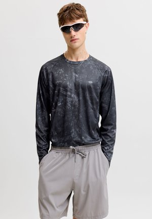 Junger Mann trägt schwarzes, gemustertes Langarmhemd, hellgraue Shorts mit Kordelzug und weiße futuristische Sonnenbrille vor einfarbigem Hintergrund.