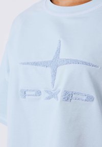 Lichtblauwe katoenen t-shirt met een textuur, geborduurd sterrenlogo en de merkinitialen "PXP" in een zacht, contrasterend materiaal.