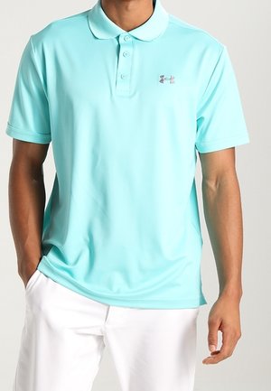 Man draagt lichtblauw poloshirt met korte mouwen met Under Armour-logo en witte broek, één hand in de zak, tegen een effen achtergrond.