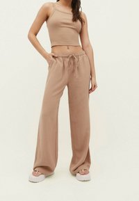 Vrouw in een beige mouwloos crop-top met bijpassende wijde spijkerbroek met trekkoord en witte platform sandalen, staande tegen een lichte achtergrond.