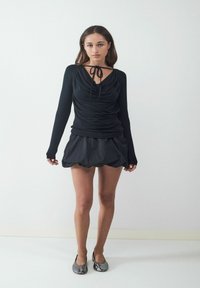 Frau steht gegen eine weiße Wand und trägt ein gerafftes schwarzes langärmliges Oberteil mit Halsband, einen schwarzen Puffrock und metallische Ballerinas.