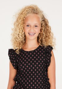 Abito nero con una texture particolare e spalle a ruffles, caratterizzato da un motivo geometrico rosa. La modella ha capelli biondi ricci e indossa l'apparecchio.