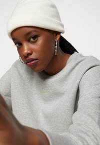 Gray sweatshirt with gold Nike logo, paired with a white knit beanie. Features include braided hair and silver spiral earrings.