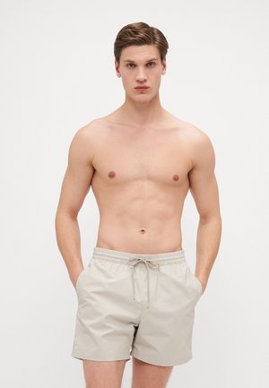Shorts de bain beiges avec une taille élastique et un cordon de serrage. Les shorts disposent de poches latérales et d'une texture de tissu lisse et léger.