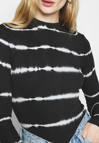 Langærmet top i sort med hvide tie-dye striber. Har ribbet tekstur og høj hals, kombineret med lyseblå denimjeans.