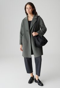 Grau wollener Mantel mit Reverskragen, zwei Knöpfen und seitlichen Taschen, kombiniert mit schwarzen, cropped Jeans und Loafers, hält eine schwarze Tasche.