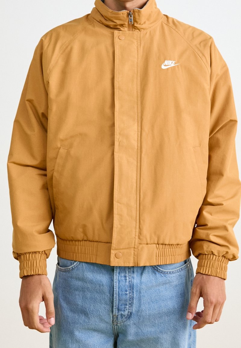 Ljusbrun Nike-jacka med hög krage, dragkedjestängning, elastiska ärmslut och sidofickor; slät tyg och minimalistisk logodetalj.