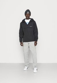 Black lightweight pullover jacket with a front pocket, paired with gray jeans and white sneakers. The model also wears a black beanie.