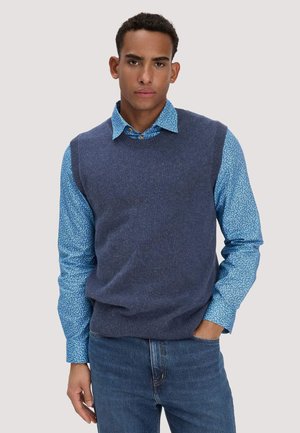 Mann trägt blau gemustertes Hemd mit Knöpfen, dunkelblaue gestrickte ärmellose Weste und blaue Jeans mit einer Hand in der Hosentasche.