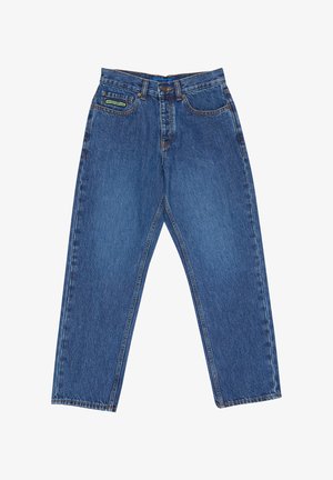 Mladá osoba s kučeravými vlasmi oblečená v bielom tričku s potlačou, modrých džínsach a čierno-bielych teniskách stojaca pred jednofarebným pozadím.