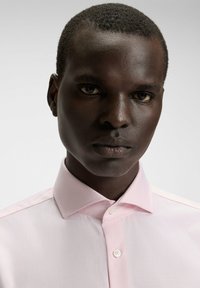 Jasnoróżowa koszula w kratkę z rozłożystym kołnierzykiem i białymi guzikami, zaprezentowana na modelce o ciemnej karnacji na neutralnym tle.