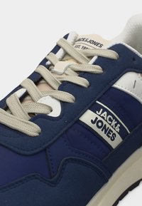 Μπλε και λευκό αθλητικό παπούτσι Jack & Jones με μπεζ κορδόνια, εμφανείς ραφές και λογότυπο στο πλάι και στη γλώσσα.