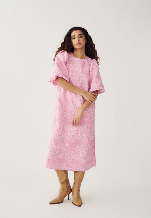 Vrouw met krullend haar, gekleed in een roze midi-jurk met een bloemenstructuur, pofmouwen en beige laarzen met hoge hak, tegen een effen achtergrond.