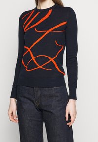 Navy sweater with orange abstract patterns, featuring a ribbed crew neck and cuffs. Soft knit fabric, fitted style, worn with dark denim.