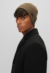 Berretto a coste marrone con una texture liscia. Indossato con un cappotto nero, mostra una vestibilità aderente e un'etichetta del logo discreta sul lato.