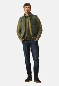 Green fleece vest over a green shirt, dark jeans, and black sneakers. The vest features a pocket and a textured surface.