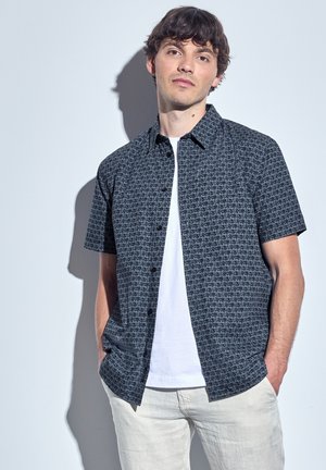 Korte mouwen button-up shirt in zwart en grijs met een geometrisch patroon, gecombineerd met een witte T-shirt met ronde hals en lichtgrijze broek.