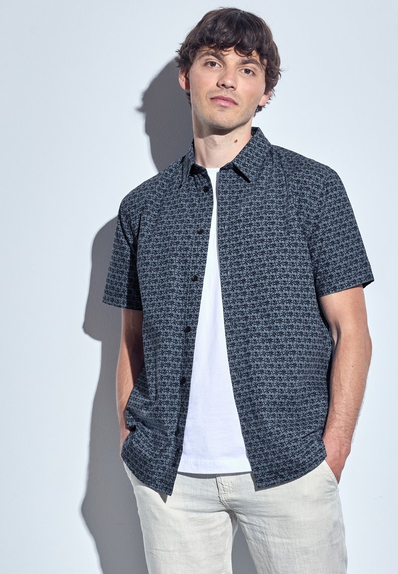 Short-sleeve button-up shirt in black and gray with a geometric pattern, paired with a white crew-neck t-shirt and light gray pants.