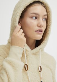 Krämfärgad fleecetröja med en texturerad, fluffig yta, en huva med dragsko och beige ribbade ärmslut med bruna hårdvaruaccenter.