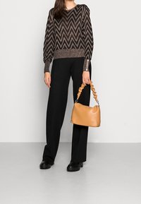 Kobieta nosząca czarne szerokie spodnie, sweter w czarno-brązowy zygzak, czarne masywne buty, trzymająca jasnobrązową torebkę z marszczonym uchwytem.