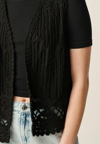 Vest negru din croșetat, cu modele intricate, design cu fața deschisă și margine ondulată, purtat deasupra unei bluze negre cu benzi verticale și a unor pantaloni scurți din denim deschis.