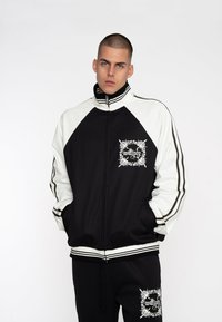 Sort og hvid zip-up jakke med raglanærmer, der har et grafisk design på brystet og stribede detaljer langs ærmerne.