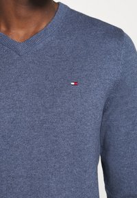 Niebieski dziergany sweter z dekoltem w serek, z małym, wielokolorowym logo na piersi. Faktura gładka, z prążkowanym wykończeniem przy szyi.