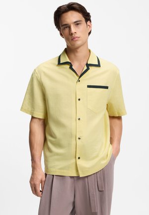 Jeune homme portant une chemise jaune à manches courtes avec une bordure noire sur le col et la poche, assortie à un pantalon gris ample.