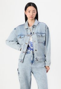 Chaqueta de mezclilla azul claro con un diseño clásico, dos bolsillos en el pecho y cierres de botón de metal. La tela muestra una textura desgastada.