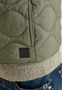 Olivengroene gewatteerde vest met ronde patronen, voorzien van een zwart logo-label en een gestructureerde crèmekleurige gebreide zoom, gecombineerd met blauwe spijkerbroek.