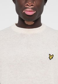 Pull en tricot beige clair avec un col rond, comportant un petit logo brodé noir et jaune sur le côté gauche de la poitrine.