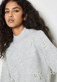 Hellgrauer Pullover aus weichem Material, verziert mit aufwendigen, mit Perlen geschmückten Schleifen. Rundhalsausschnitt, ribbelte Bündchen und Saum.