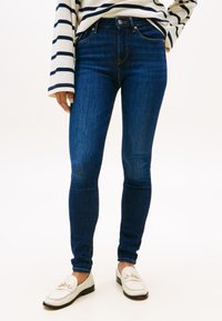 Isik, kes kannab tumesiniseid kitsaid teksaseid, valget ja mereväe triibulist pika varrukaga särki ning valgeid kuldsete detailidega mokasiine.