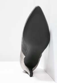 Scarpa con tacco alto grigia con punta affusolata, caratterizzata da una tomaia in pelle liscia e una suola in gomma testurizzata, inclinata su uno sfondo bianco.