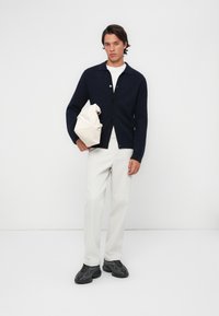 Cardigan tricotat de culoare bleumarin cu guler ascuțit, nasturi negri, tricou alb, pantaloni crem și adidași negri. Ținând o geantă crem.