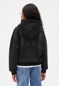 Persona ar gariem tumšiem matiem un caurspīdīgām brillēm, valkājot melnu kapuci ar spīdīgām burtiem un zilas džinsu bikses, skatās prom no kameras.