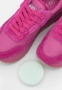 Baskets rose vif en mesh et suède, avec des semelles épaisses et des lacets ronds. Une éponge circulaire vert clair est à proximité.