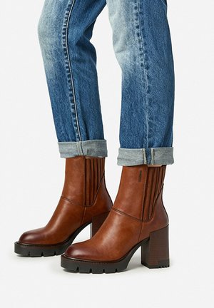 Botines de cuero marrón con tacones gruesos, usados con jeans de mezclilla azul remangados sobre un fondo blanco liso.