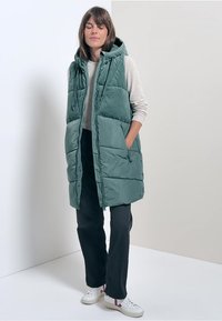 Zielona, ocieplana kamizelka bez rękawów z kapturem, z dwoma bocznymi kieszeniami i pikowanym wzorem, w połączeniu z jasnym swetrem i czarnymi spodniami.