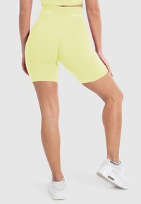 Geel sportieve fietsshorts met een hoge tailleband, gemaakt van rekbare stof, met een logo aan de bovenkant. Gdragend met witte sneakers.