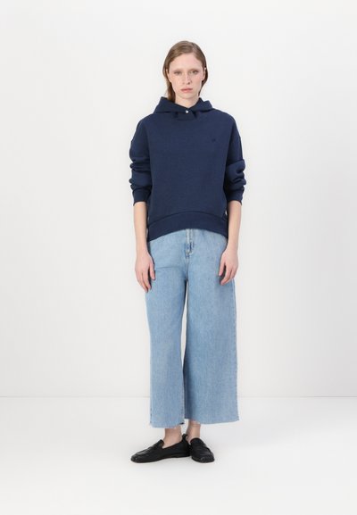 Sweatshirt à capuche bleu marine avec une poche avant, associé à un jean large bleu clair. Le modèle porte des chaussures noires à enfiler. Fond uni.